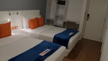 Habitación de hotel con dos camas, cada una con una manta azul y almohadas naranjas. Se observa un armario blanco y una mesita con una lámpara.