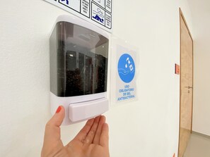 Una mano presionando un dispensador de gel antibacterial instalado en la pared, con un letrero que indica su uso.