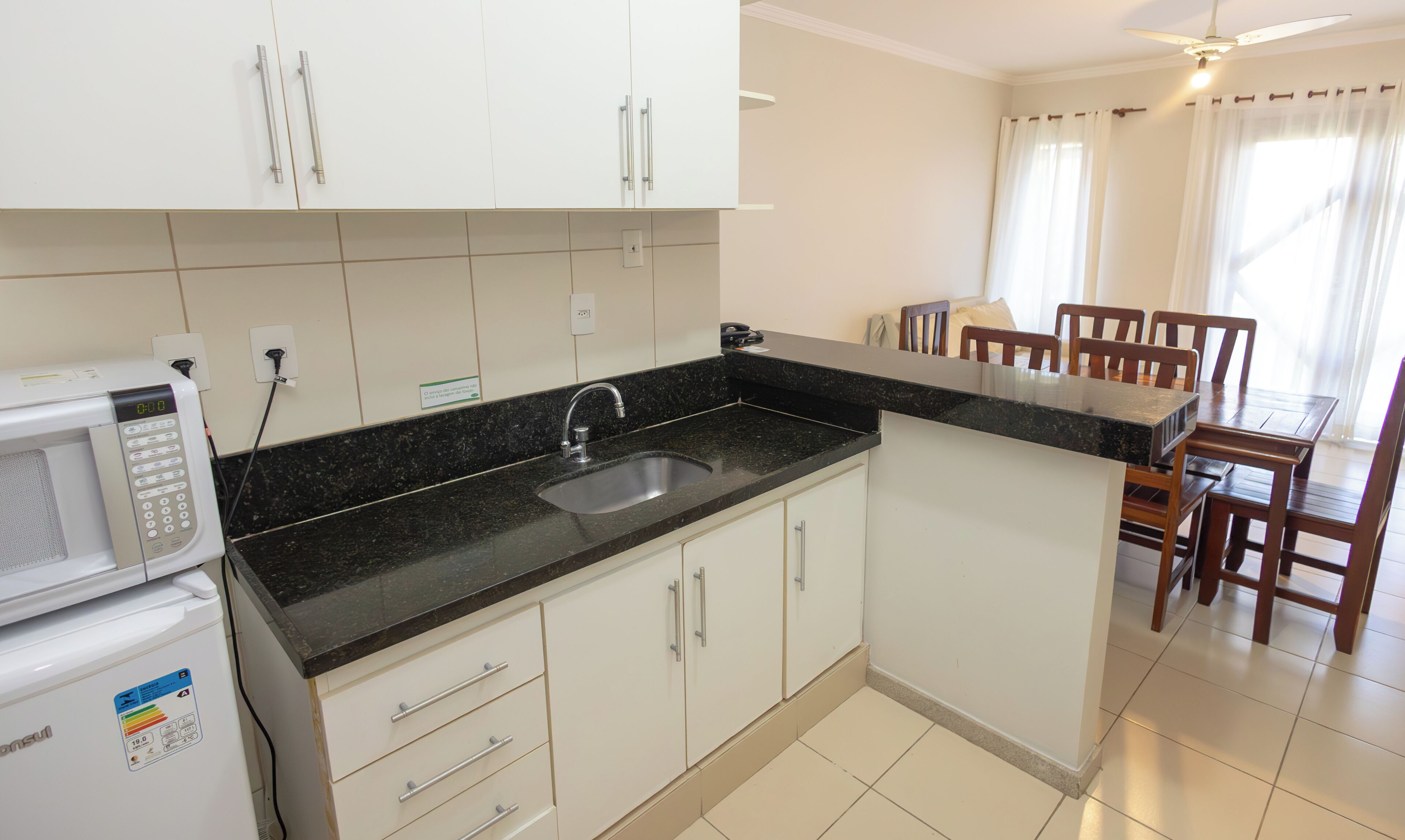A compact kitchen with white cabinets, a black countertop, a built-in sink, and a microwave. There is a dining area with wooden chairs and a table.