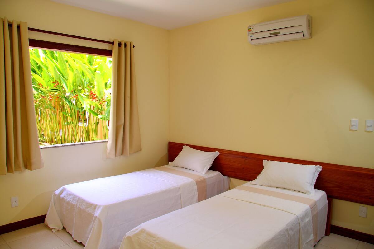 A room with two single beds, beige walls, a window with curtains, and an air conditioning unit.