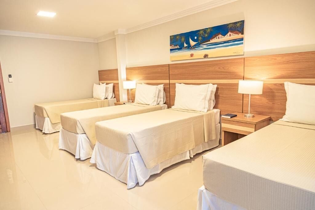 Ein Hotelzimmer mit drei Einzelbetten, jeweils mit weißer Bettdecke und passenden Kissen. Es gibt ein Holz-Kopfteil, ein Bild mit einer Strandansicht und einen Nachttisch mit einer Lampe.