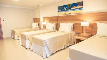 Ein Hotelzimmer mit drei Einzelbetten, jeweils mit weißer Bettdecke und passenden Kissen. Es gibt ein Holz-Kopfteil, ein Bild mit einer Strandansicht und einen Nachttisch mit einer Lampe.