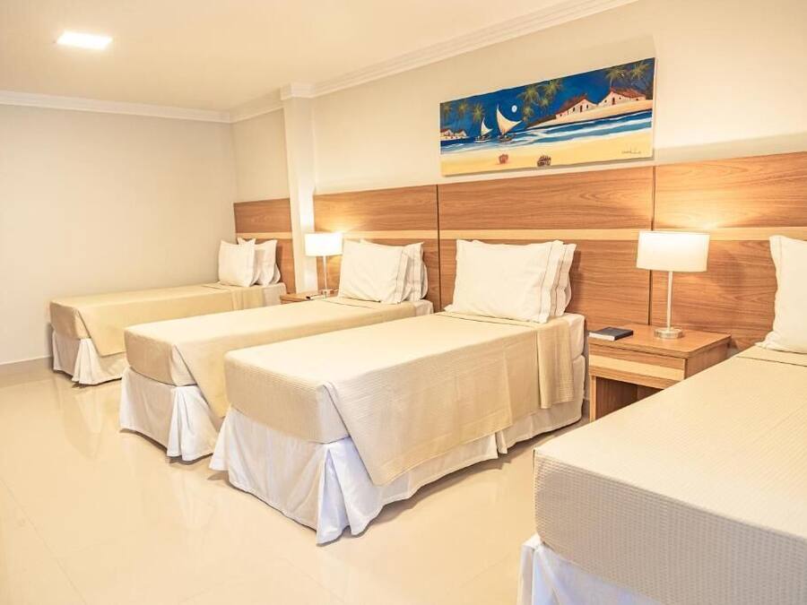 A hotel room with three single beds, each with a white bedspread and matching pillows. There is a wooden headboard, a painting of a beach scene, and a nightstand with a lamp.