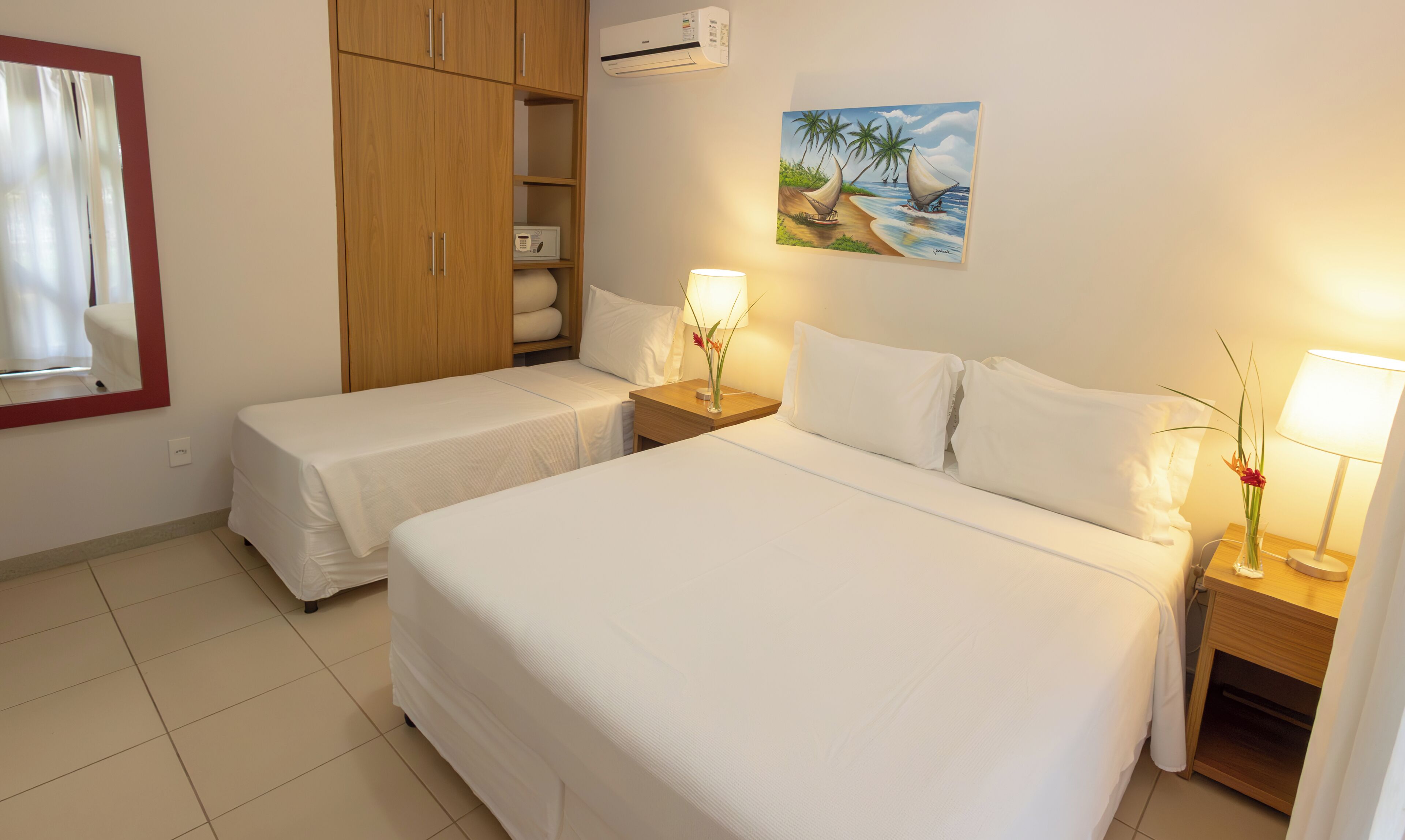 Une chambre d’hôtel avec deux lits, une armoire, un climatiseur, un tableau représentant une scène de plage et une table de chevet avec une lampe.