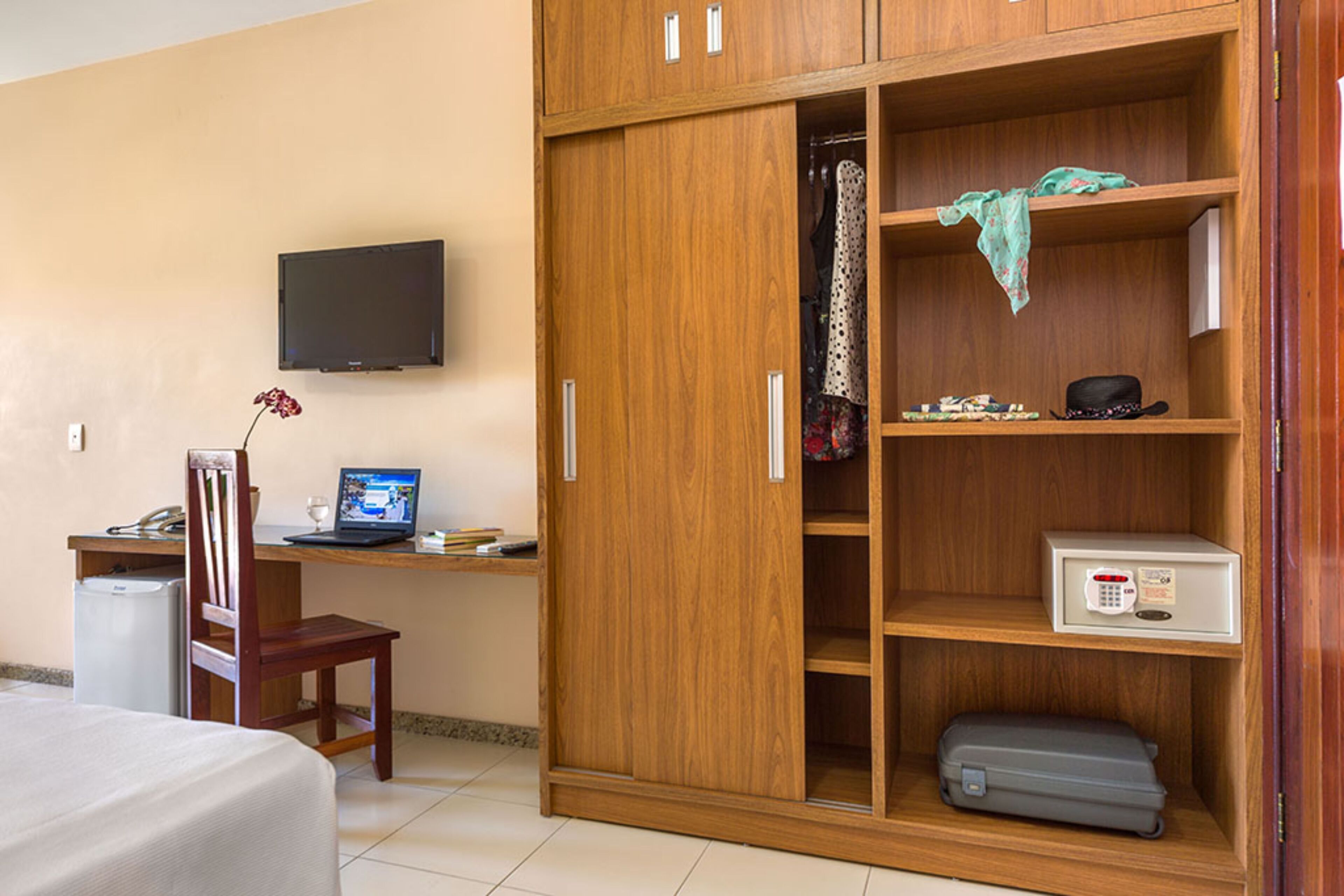 A hotel room with a wooden wardrobe, a desk with a computer, a television mounted on the wall, and a bed.