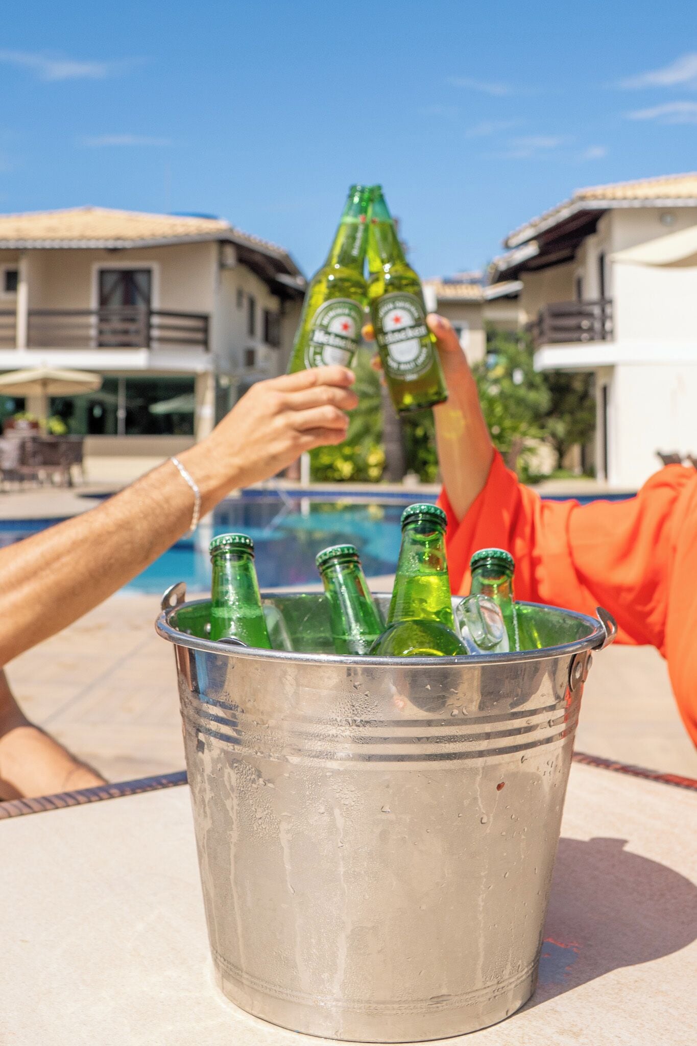 A bucket with several green bottles, likely containing beer, placed on a table near a poolside area with buildings in the background.