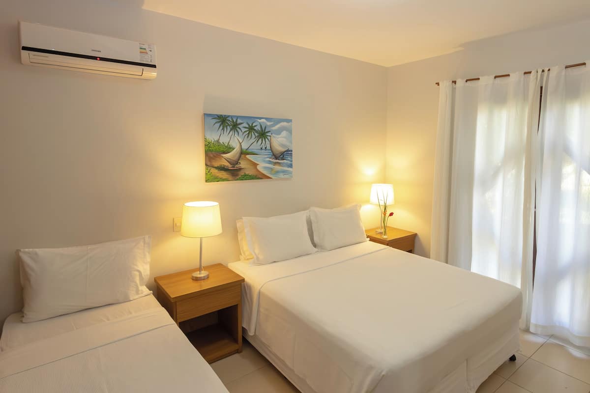 A hotel room with two beds, a painting of a sailboat, a window with white curtains, and an air conditioning unit on the wall.