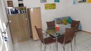 Un coin repas avec une table en verre, des chaises en osier, un réfrigérateur et un meuble contenant des bouteilles.