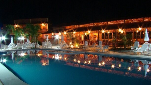 Un espace piscine aménagé pour la nuit, avec des chaises longues et des parasols, un bâtiment à l’éclairage chaleureux et une piscine aux eaux claires qui reflète les lumières.