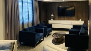A modern living room with a flat-screen TV, dark blue sofas, a white console table, and a large window with blinds. - Hotel Kennedy (Sao Jose)