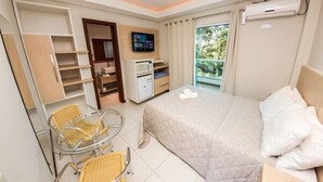 A hotel room with a bed, a TV, a minibar, a chair, and a window with a view of greenery. - Pousada Cristalmar (Bombinhas)