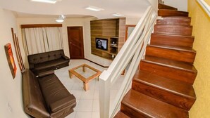 A modern living room with a sofa, a coffee table, and a TV. A wooden staircase leads to an upper floor. - Pousada Cristalmar (Bombinhas)