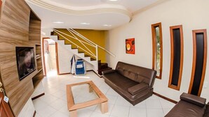 A modern living room with a brown sofa, a glass coffee table, a TV mounted on the wall, and a staircase leading to another level. - Pousada Cristalmar (Bombinhas)