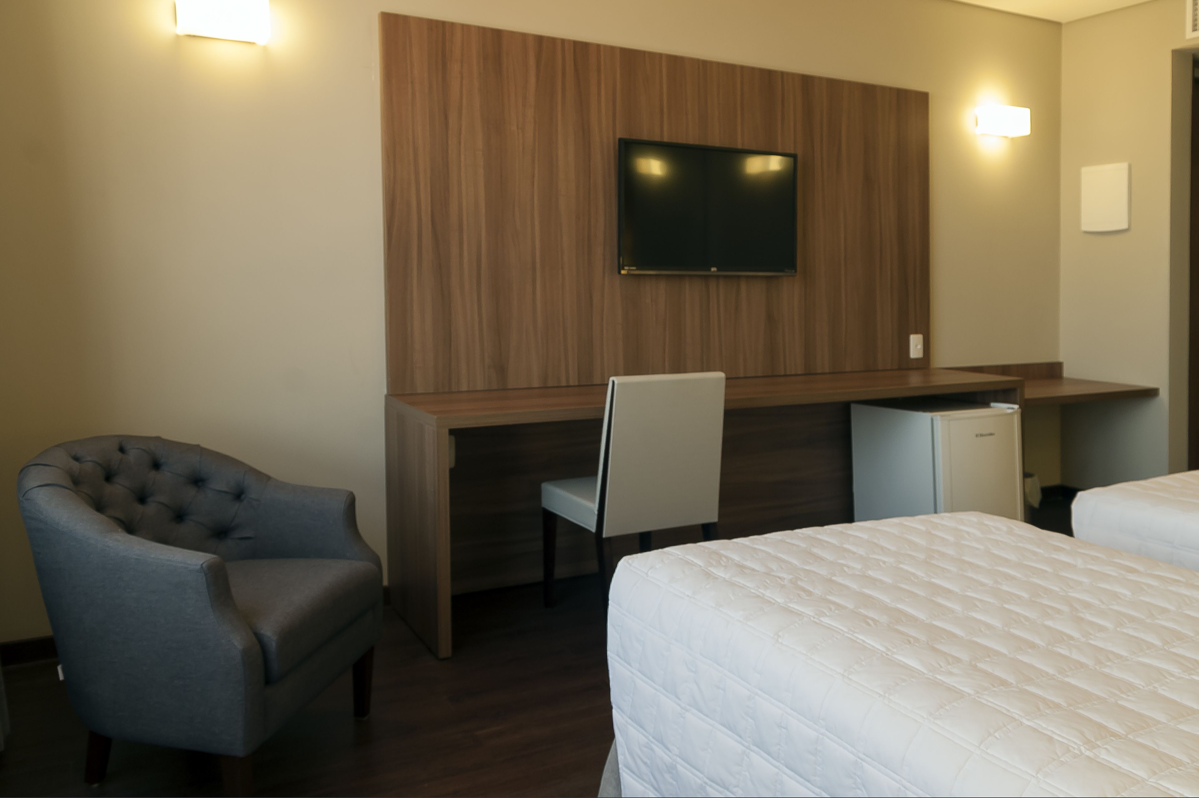Een hotelkamer met een houten bureau, een televisie, een gestofte fauteuil, een bed en een kleine koelkast.