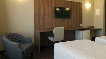 Quarto de hotel com escrivaninha de madeira, televisão, poltrona estofada, cama e um pequeno frigorífico.