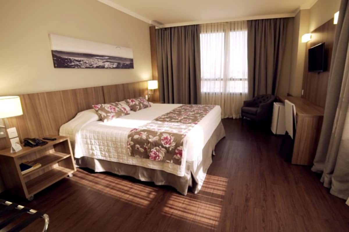 A hotel room with a large bed, a desk, a chair, a TV, and a window with curtains.