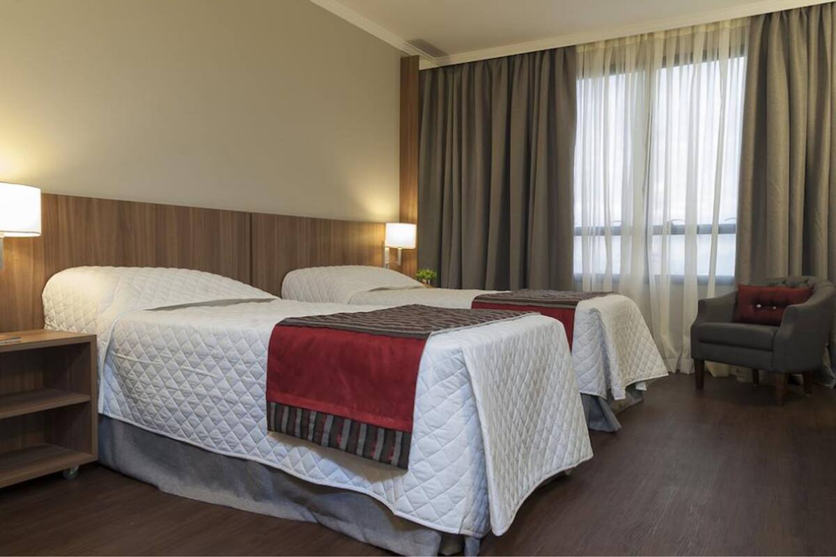 A hotel room with two beds, a wooden headboard, a desk with a lamp, a chair, and a large window with curtains.