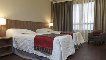 A hotel room with two beds, a wooden headboard, a desk with a lamp, a chair, and a large window with curtains.