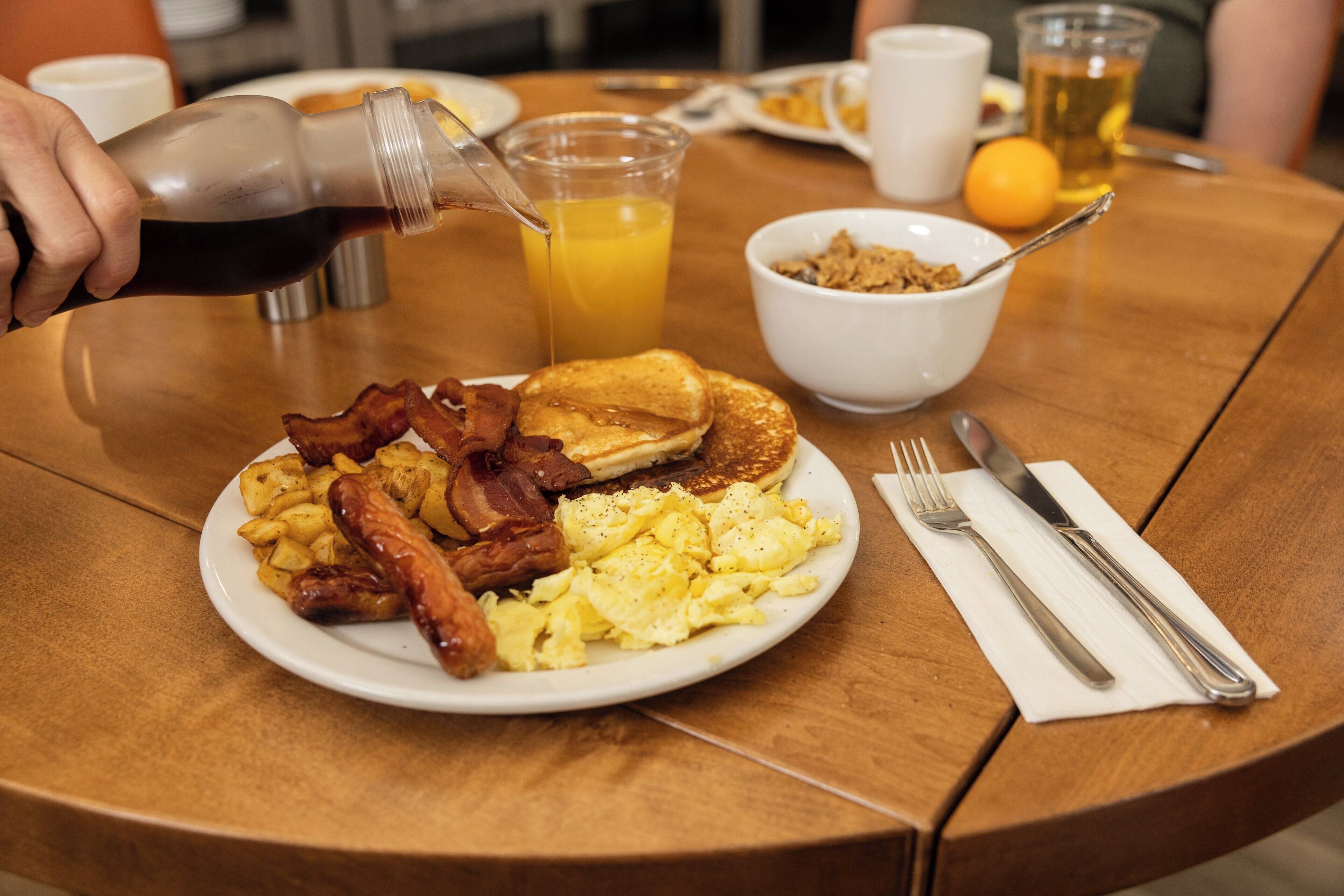 A breakfast plate with bacon, eggs, and toast, a glass of orange juice, and a bottle of syrup being poured.