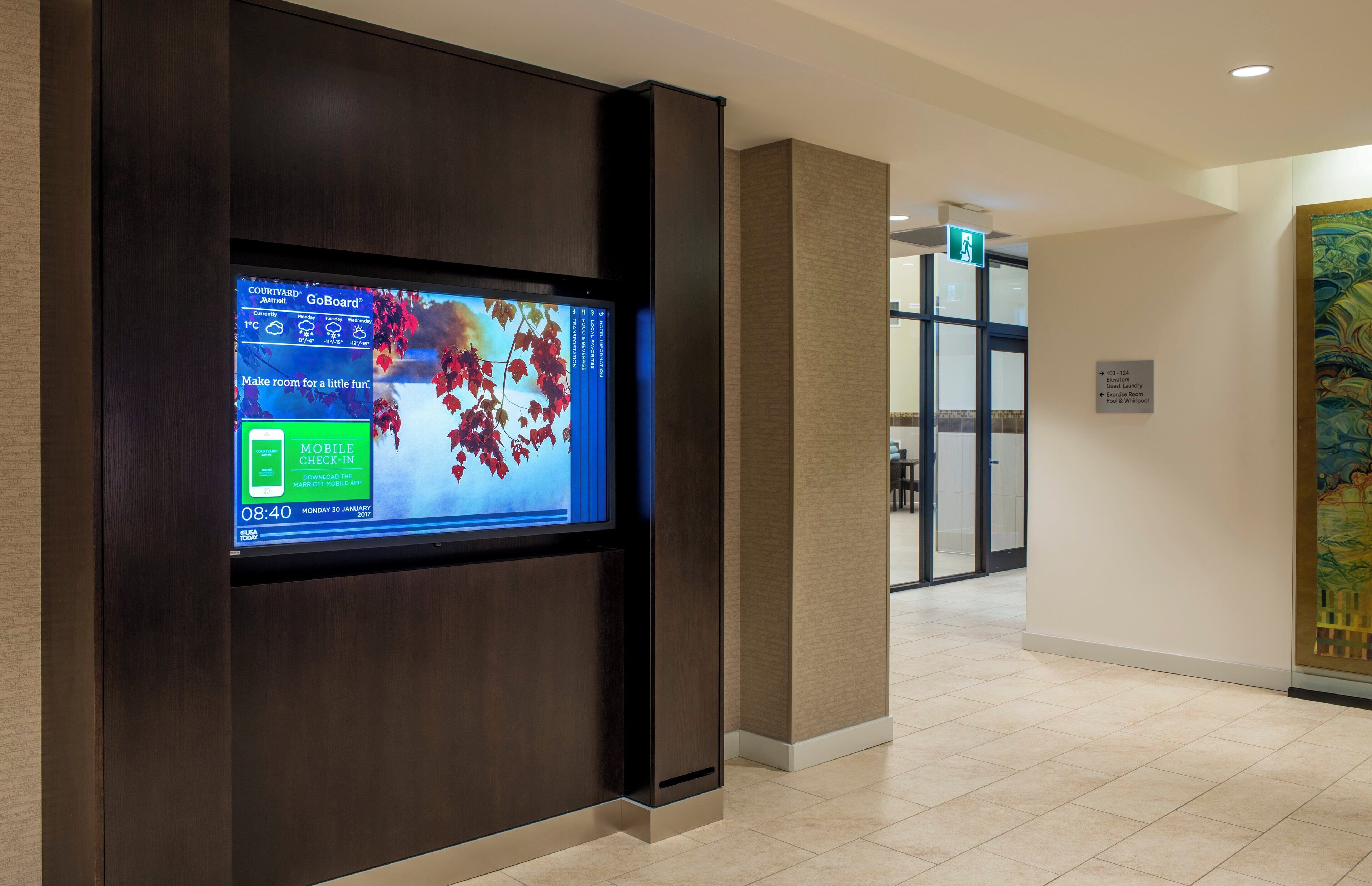 A hotel lobby with a digital display showing weather and room availability, a reception desk, and a painting on the wall.