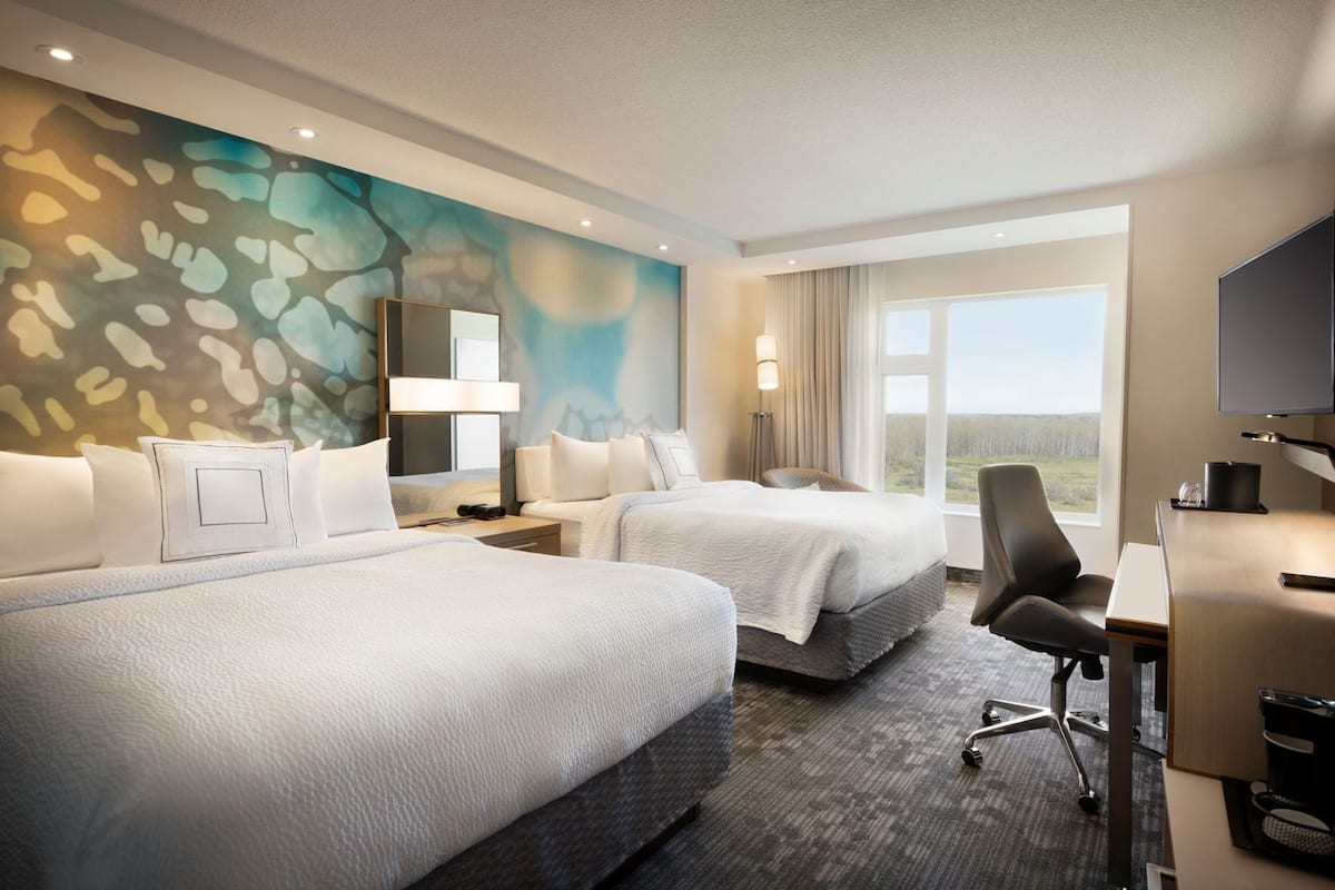 A hotel room with two beds, a desk, a chair, a TV, and a large window.