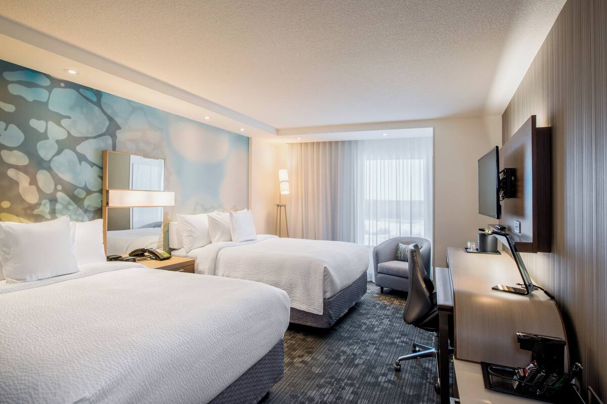 A hotel room with two beds, a desk, a chair, a TV, and a large window with curtains.