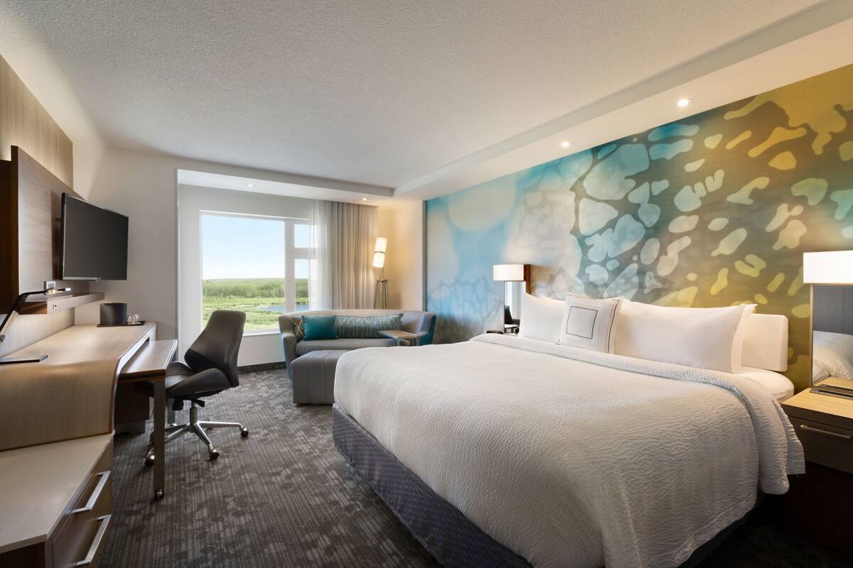 A hotel room with a bed, a desk, a chair, a sofa, a TV, and a large window.