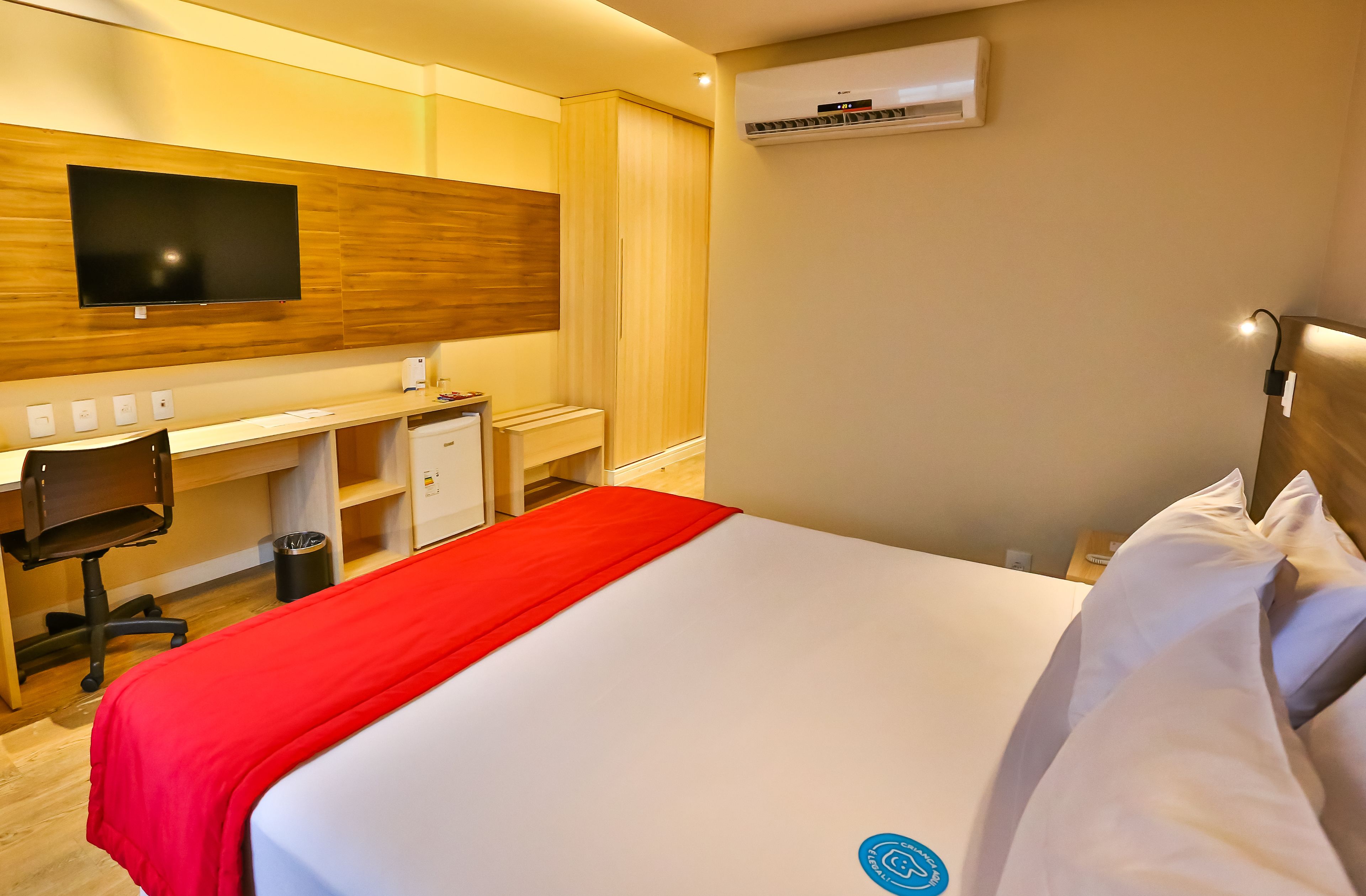 A hotel room with a bed, a desk, a chair, a TV, and an air conditioning unit.