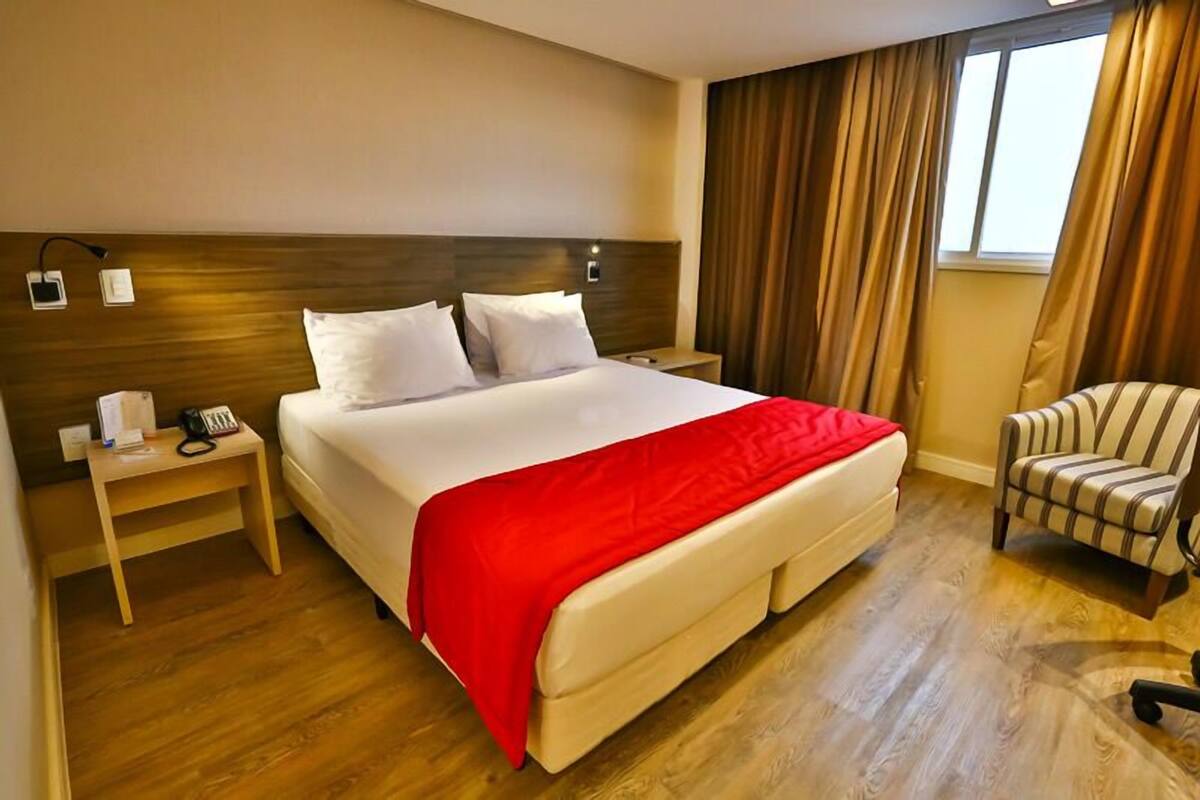 Ein Hotelzimmer mit einem großen Bett, einem Nachttisch mit Telefon, einem Sessel und einem Fenster mit Vorhängen.