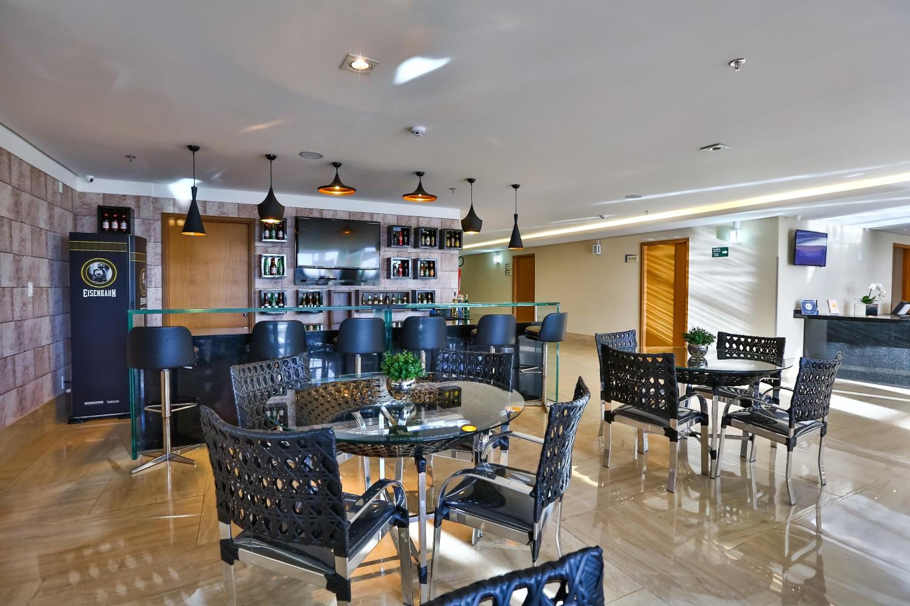 Un lobby de hotel moderno con zona de comedor de vidrio, sillas negras y un bar con estantes llenos de botellas.