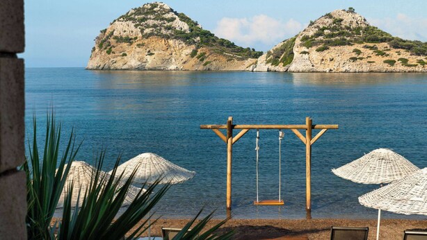 A beach with a wooden swing set and white umbrellas, overlooking calm waters and rocky hills. - Wyndham Datca Perili Bay (Datça)