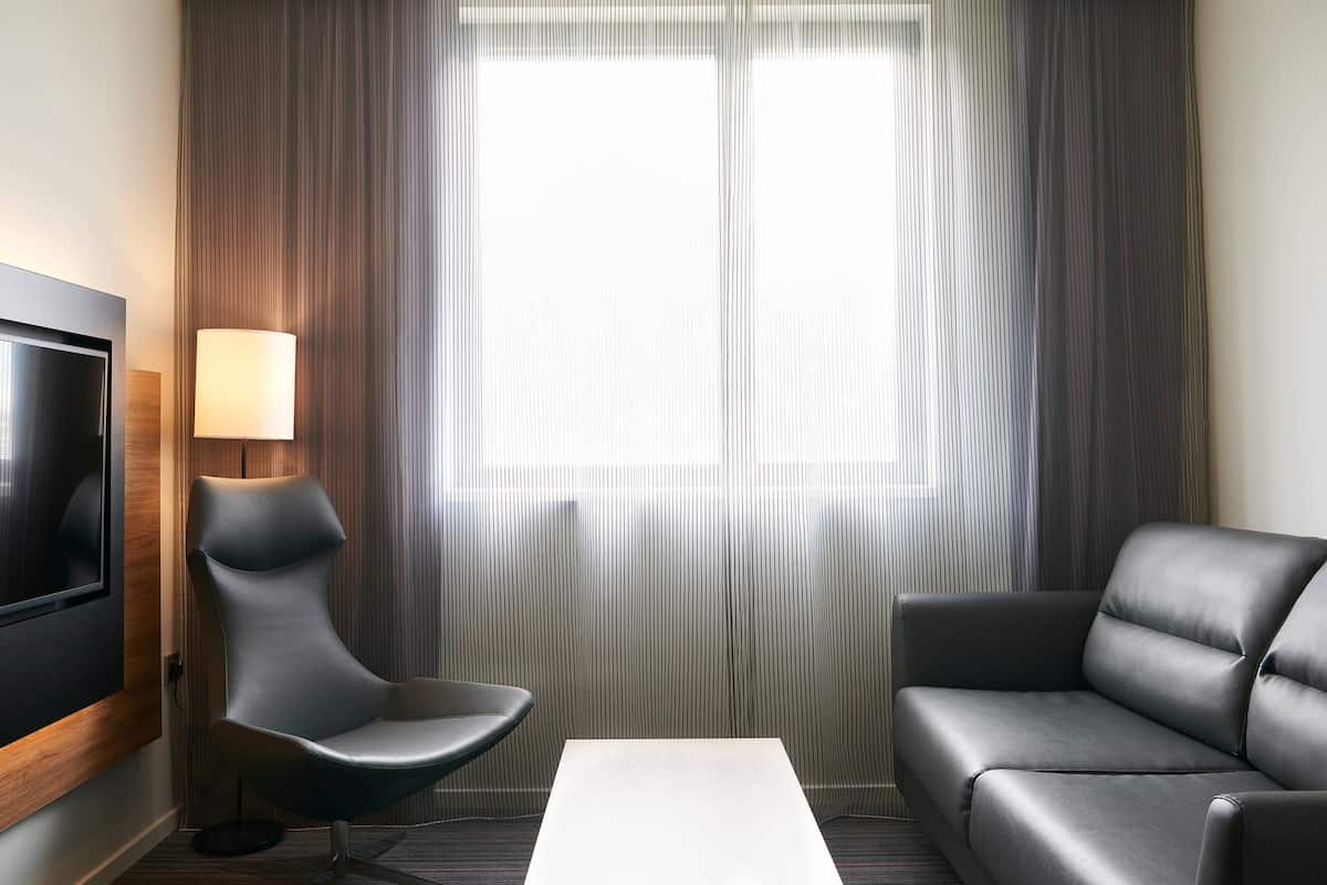 A modern hotel room with a flat-screen TV mounted on the wall, a grey sofa, a black armchair, and a small table.