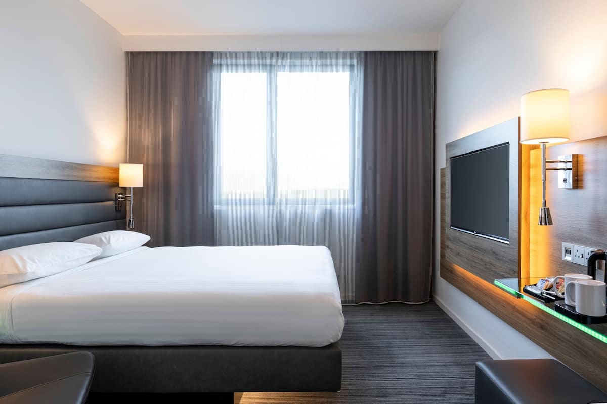 A modern hotel room with a large bed, a flat-screen TV, a bedside table with a lamp, and a view through a window with curtains.