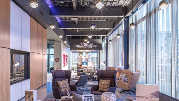 A modern lounge area with comfortable seating, a display table with books and decorative items, and large windows offering views of the city. - Moxy Aberdeen Airport (Aberdeen)