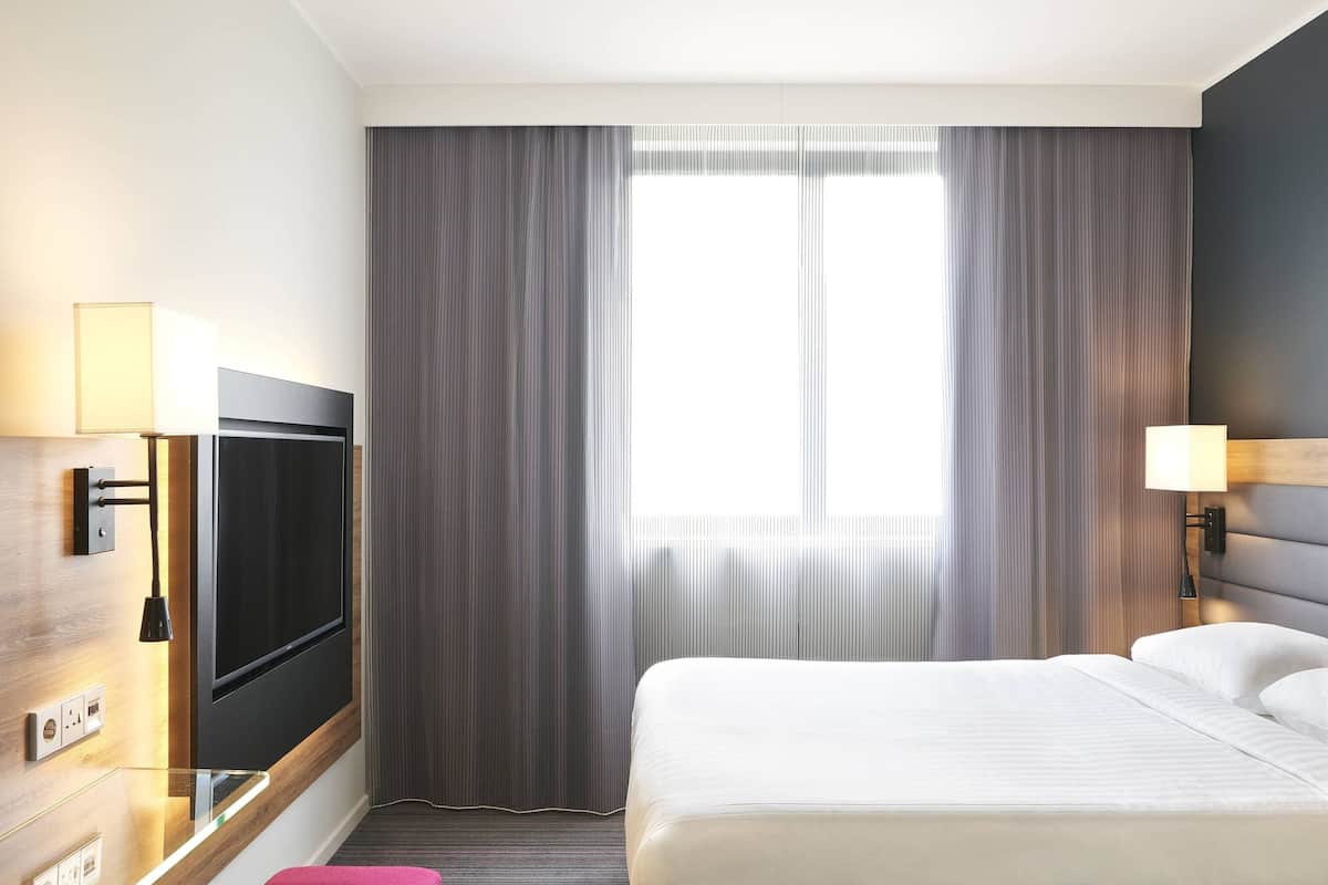 A hotel room with a bed, a TV mounted on the wall, a window with curtains, and a lamp on the nightstand.