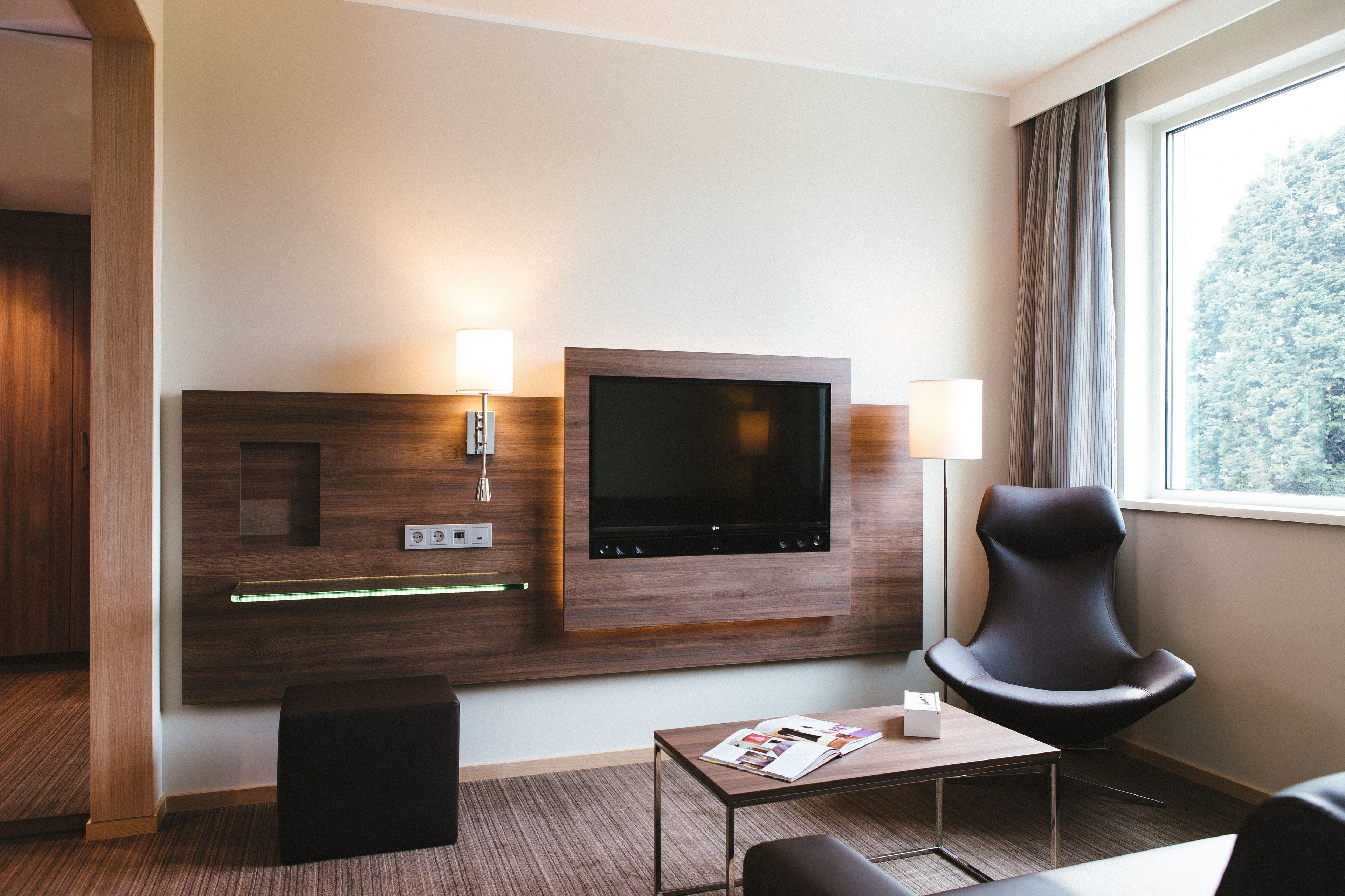 A modern hotel room with a flat-screen TV mounted on a wooden panel, a comfortable armchair, a small table with a magazine, and a window with curtains.