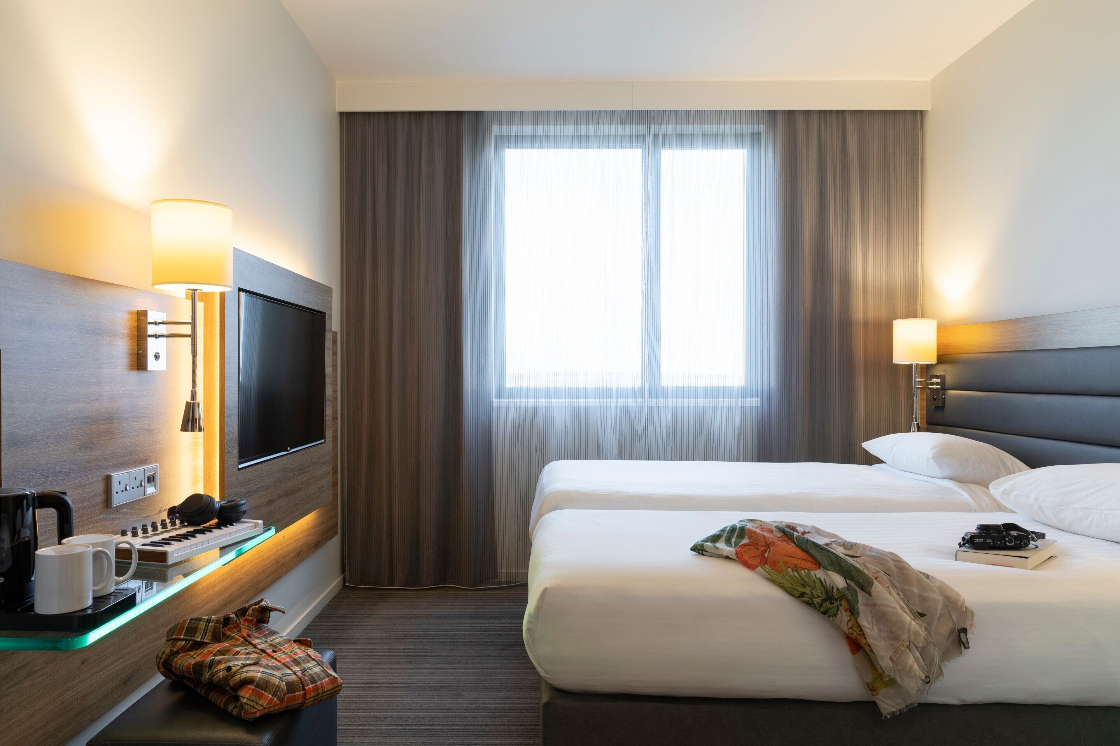 A hotel room with a bed, a TV, a desk with a keyboard and coffee maker, and a window with curtains.