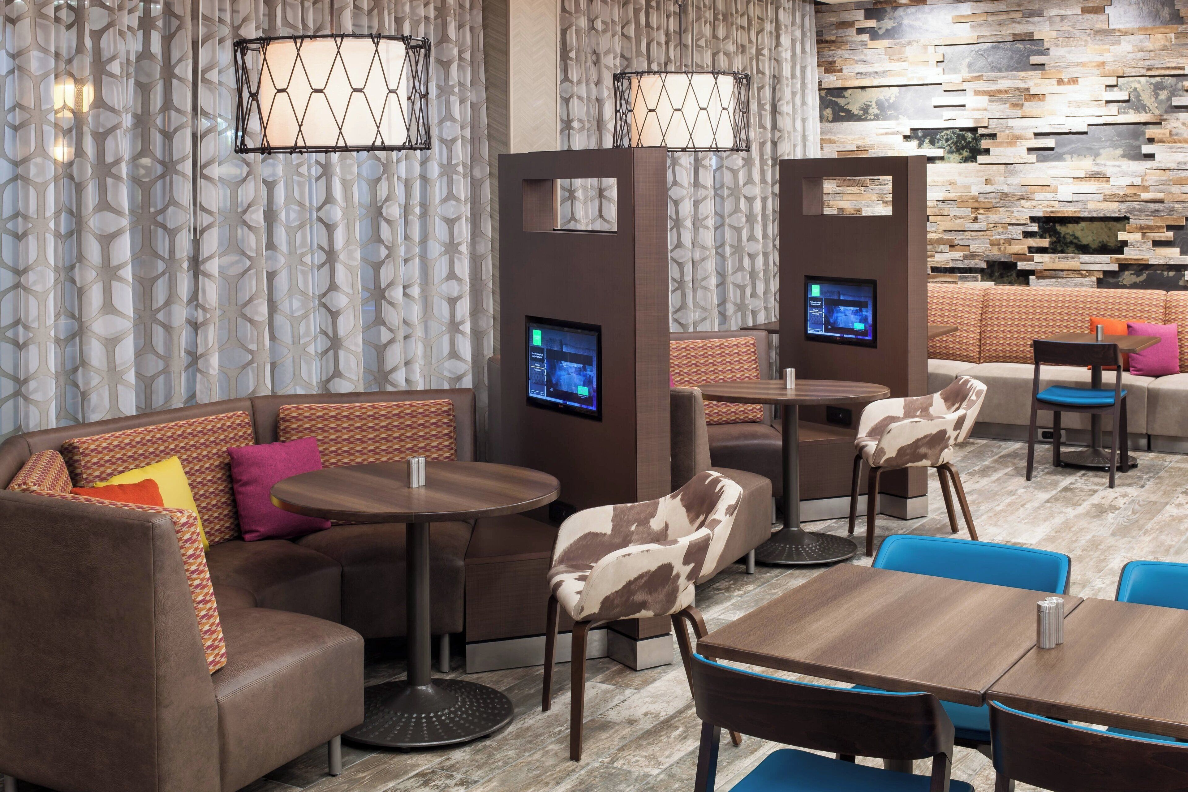 A modern lounge area with a sectional sofa, round tables, and chairs, featuring a stone wall and patterned curtains.