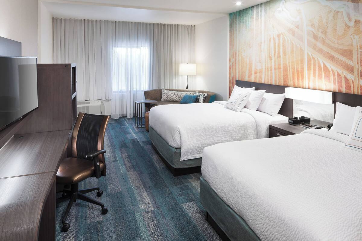 Ein Hotelzimmer mit zwei Betten, einem Schreibtisch, einem Sessel, einem Fernseher und einem Fenster mit Vorhängen.
