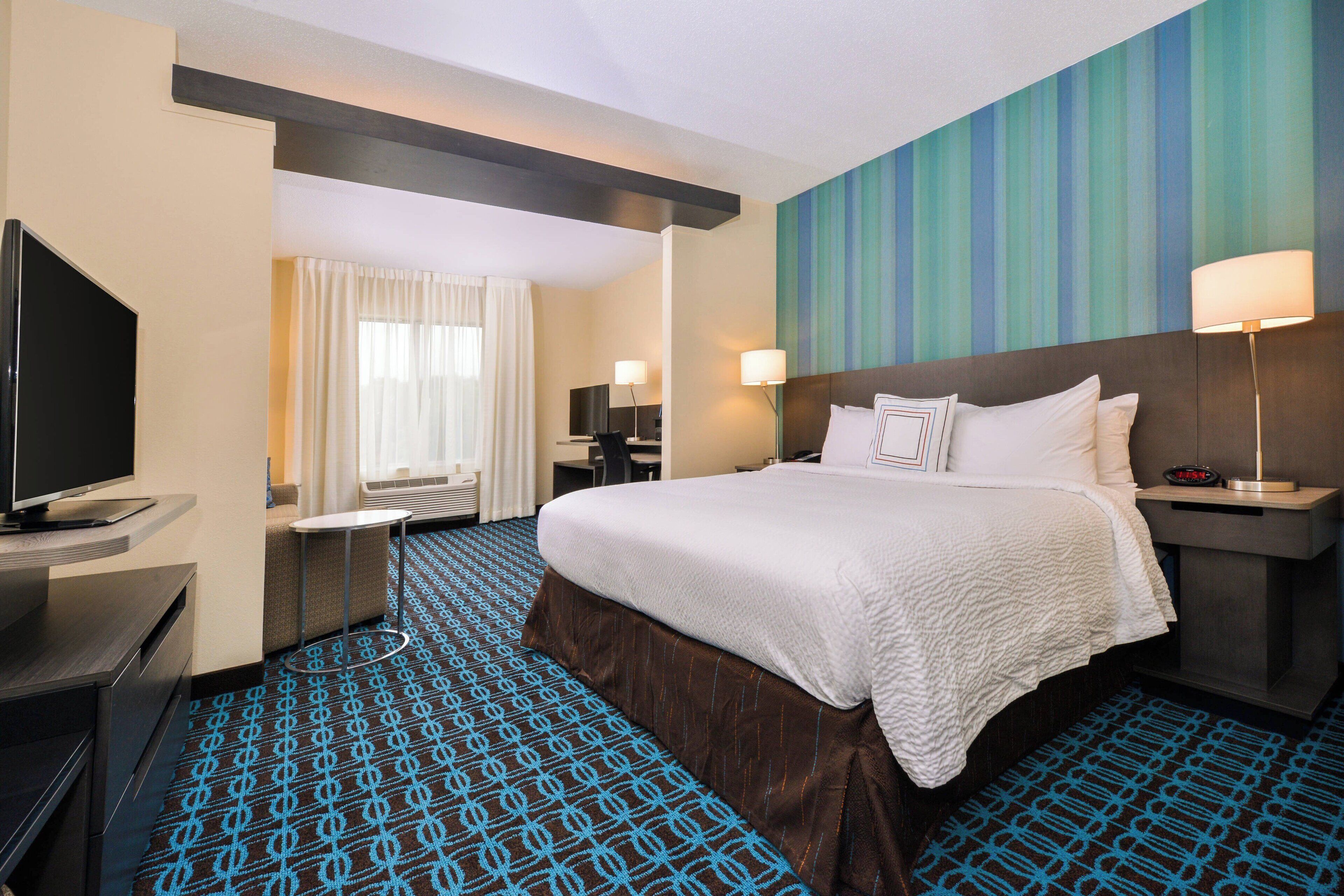A hotel room with a large bed, a flat-screen TV, a desk with a chair, and a window with curtains.
