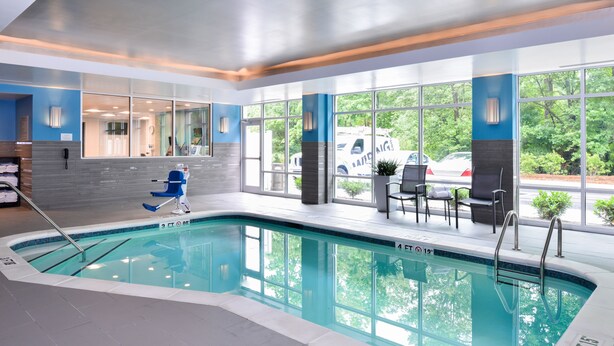 An indoor swimming pool with clear glass walls, a blue and grey tiled floor, and a view of a parked car outside. - Fairfield Inn & Suites by Marriott Raleigh Cary (Cary)