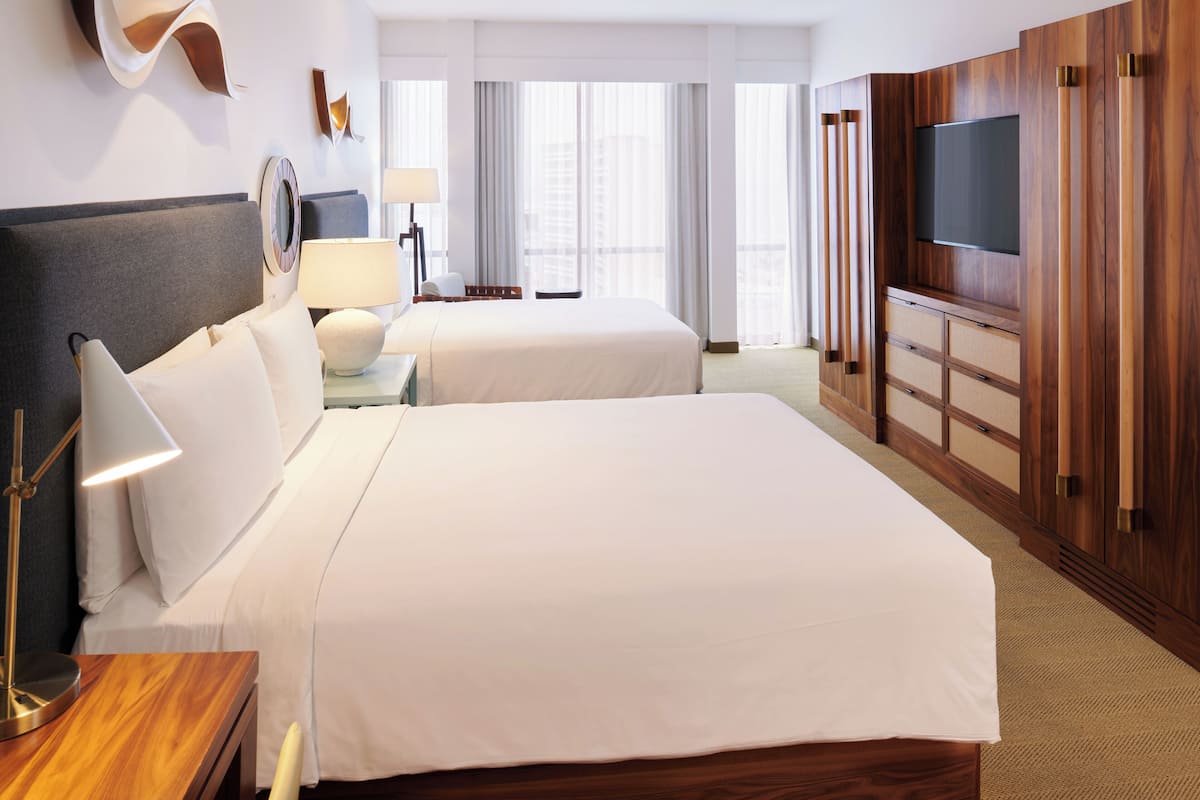 A hotel room with two beds, a TV mounted on a wooden panel, a nightstand with a lamp, and a view of the city through large windows.
