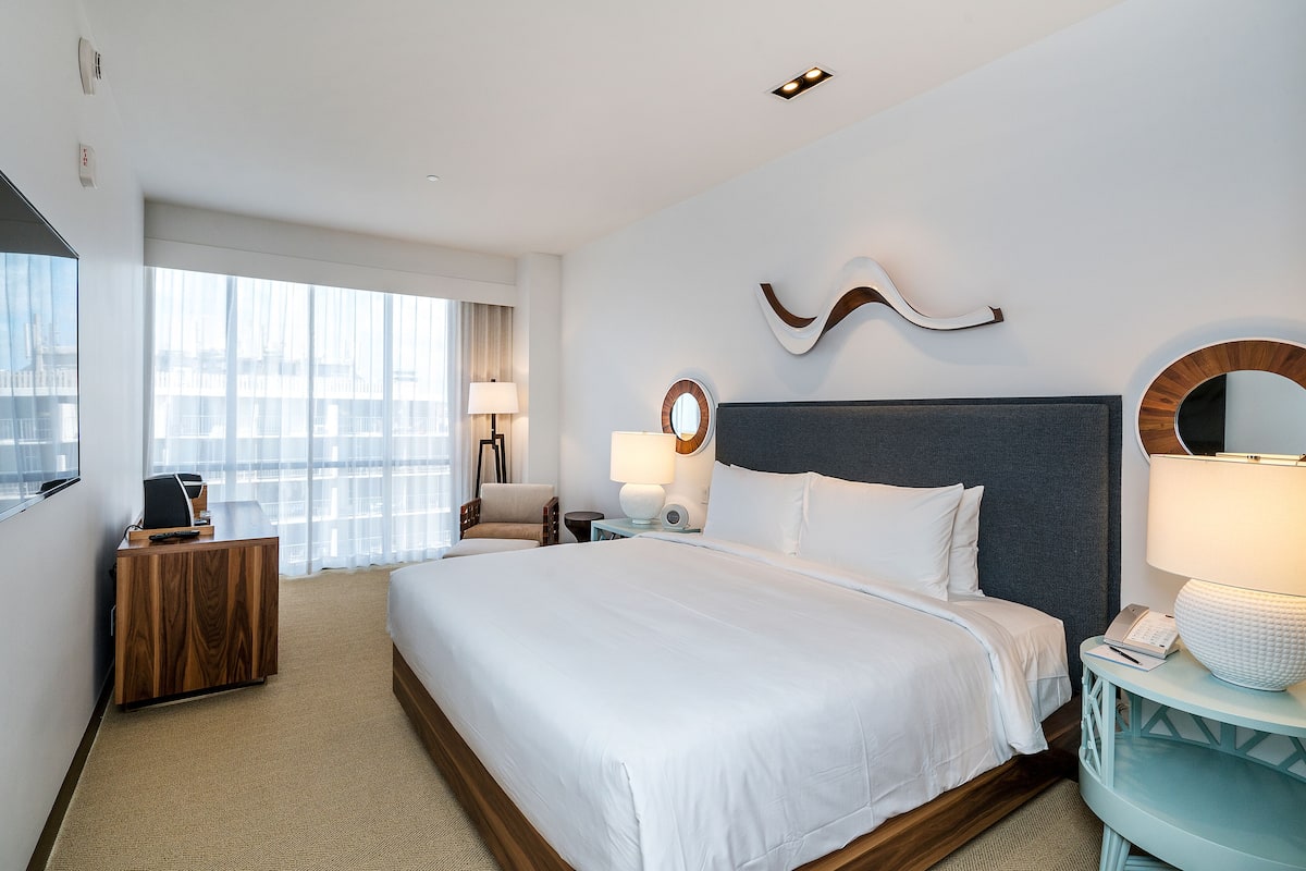 A hotel room with a large bed, a desk, a chair, a nightstand, and a window with a view of buildings.