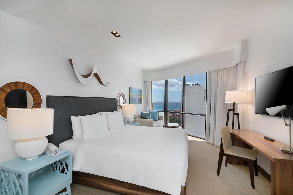 A modern hotel room with a large bed, a desk, a TV, and a balcony with a city view.