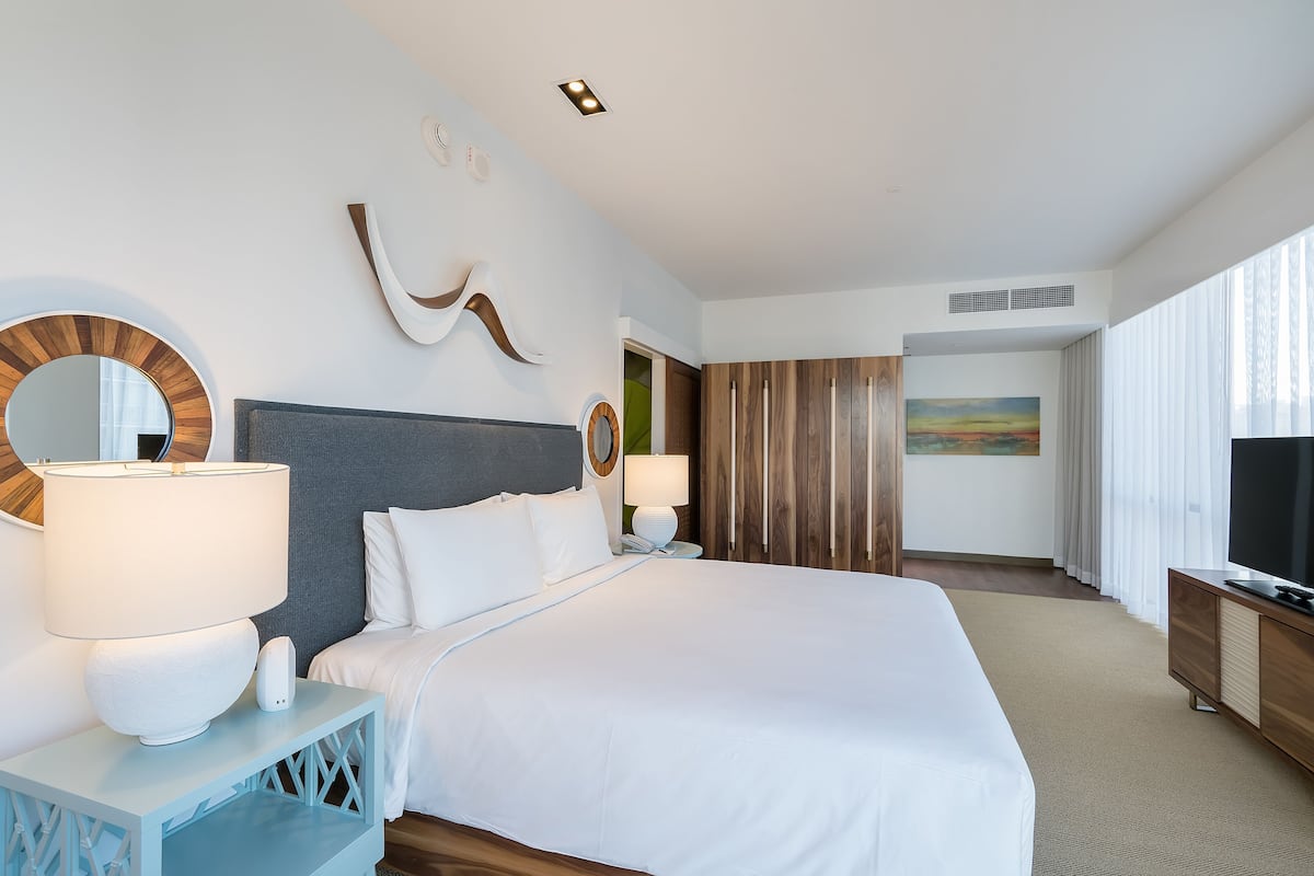 A modern hotel room with a large bed, a bedside table with a lamp, a TV, and a painting on the wall.