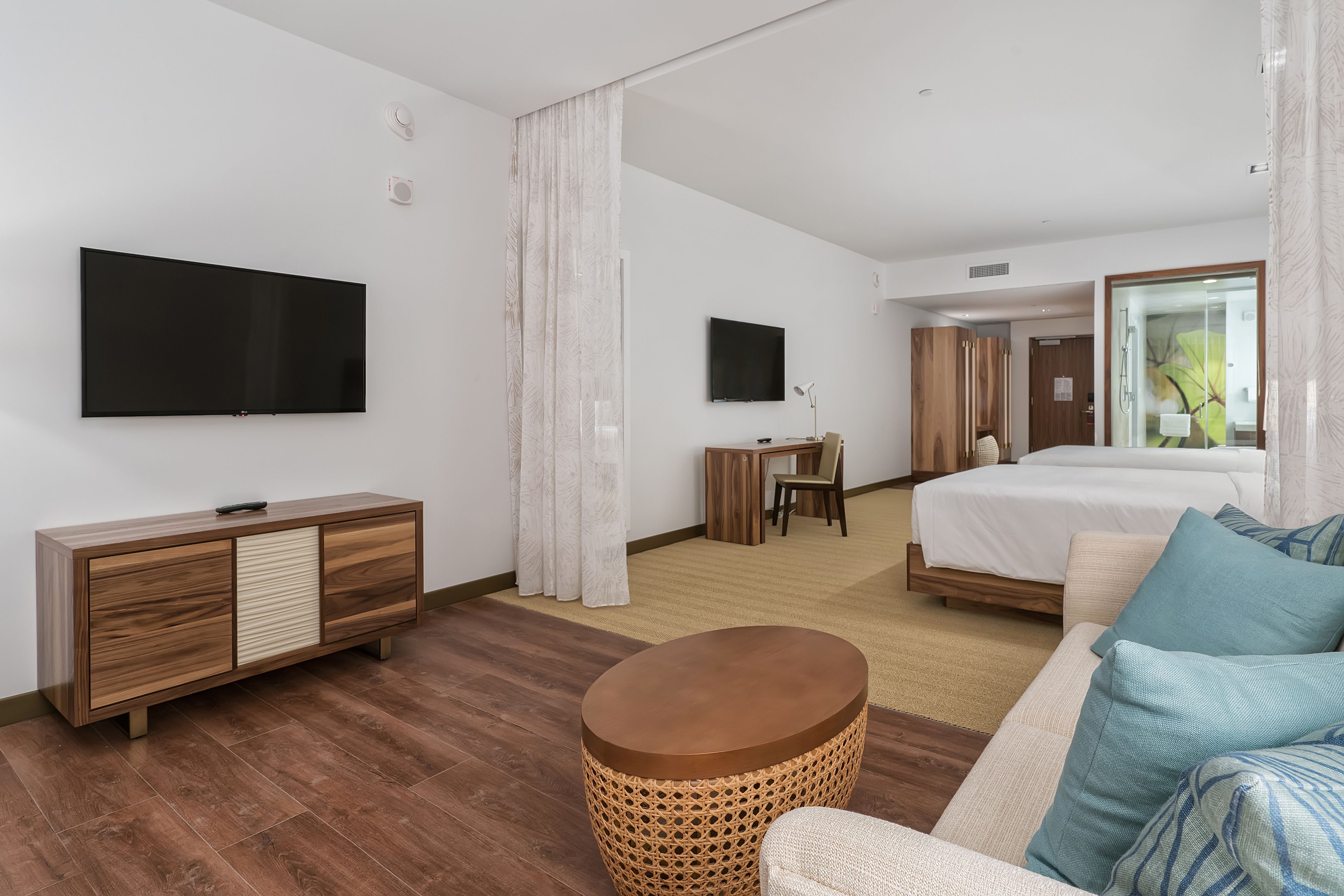 Ein modernes Hotelzimmer mit Flachbildfernseher, einem Holzkasten, einem runden Korbtisch, einer Couch mit gemusterten Kissen, einem Bett und einem Schreibtisch mit Stuhl.