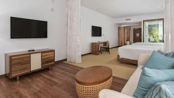 Una habitación de hotel moderna con una televisión de pantalla plana, un aparador de madera, una mesa redonda de mimbre, un sofá con cojines estampados, una cama y un escritorio con una silla.