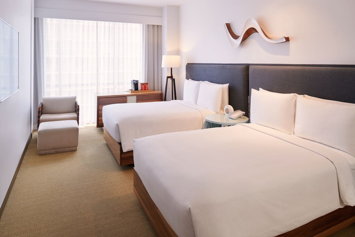 A hotel room with two beds, a chair, a bedside table with a lamp, and a large window with curtains.