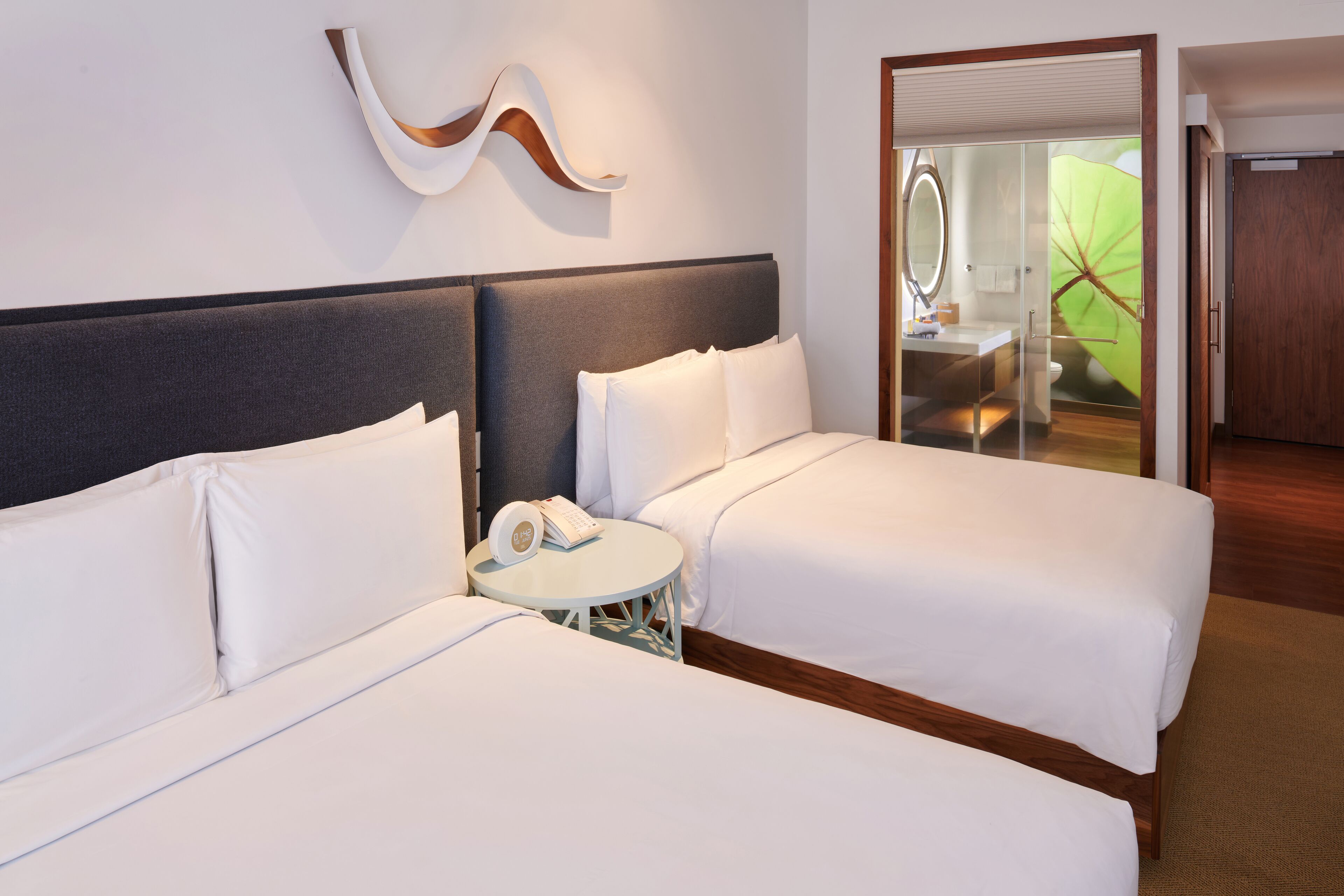 A hotel room with two beds, a small round table with a telephone, and a glass-enclosed bathroom.