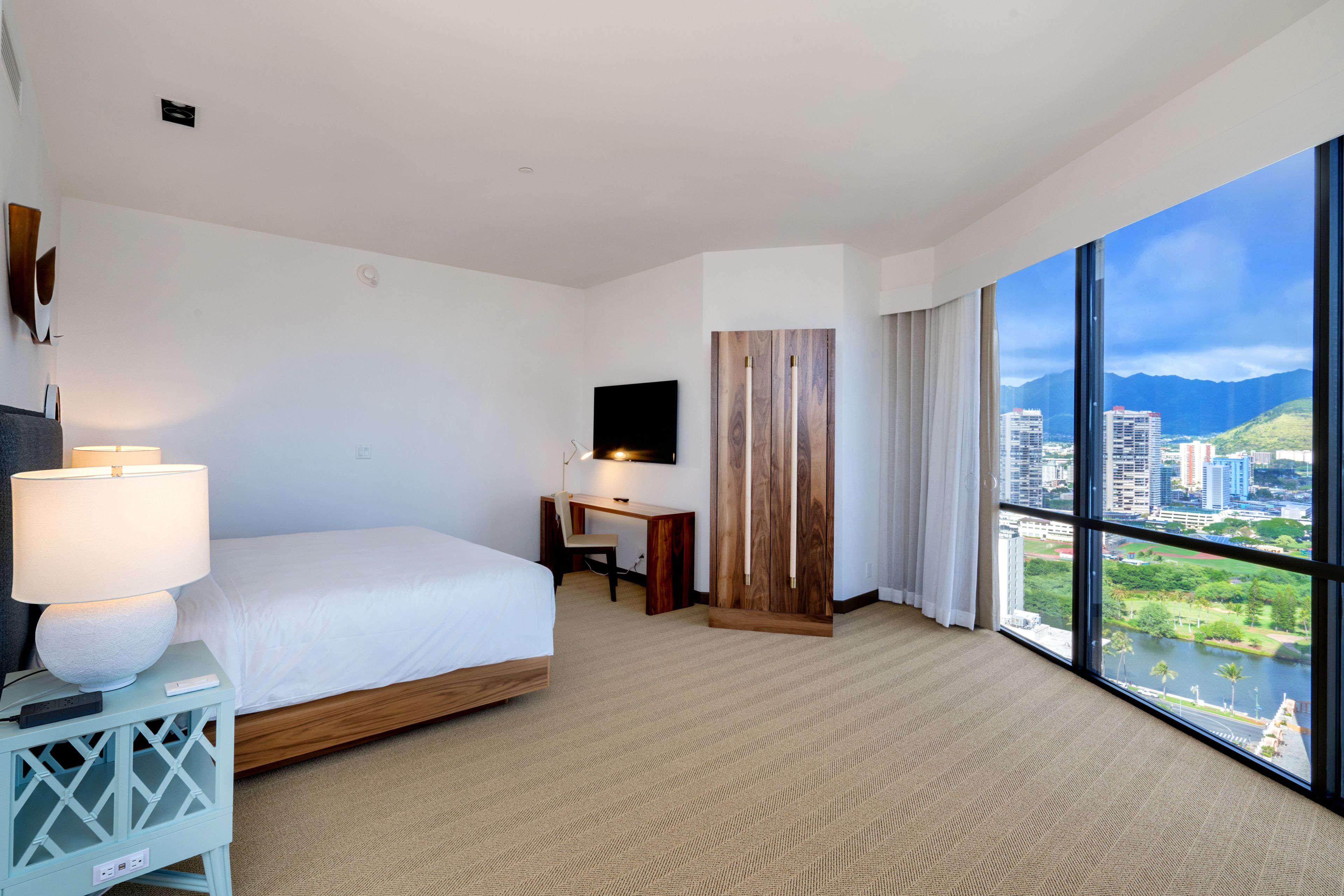 A hotel room with a bed, desk, and large windows offering a city view.
